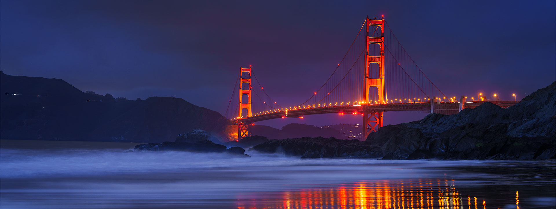 Golden Gate Bridge illuminated at night with long-exposure ocean waves and reflections