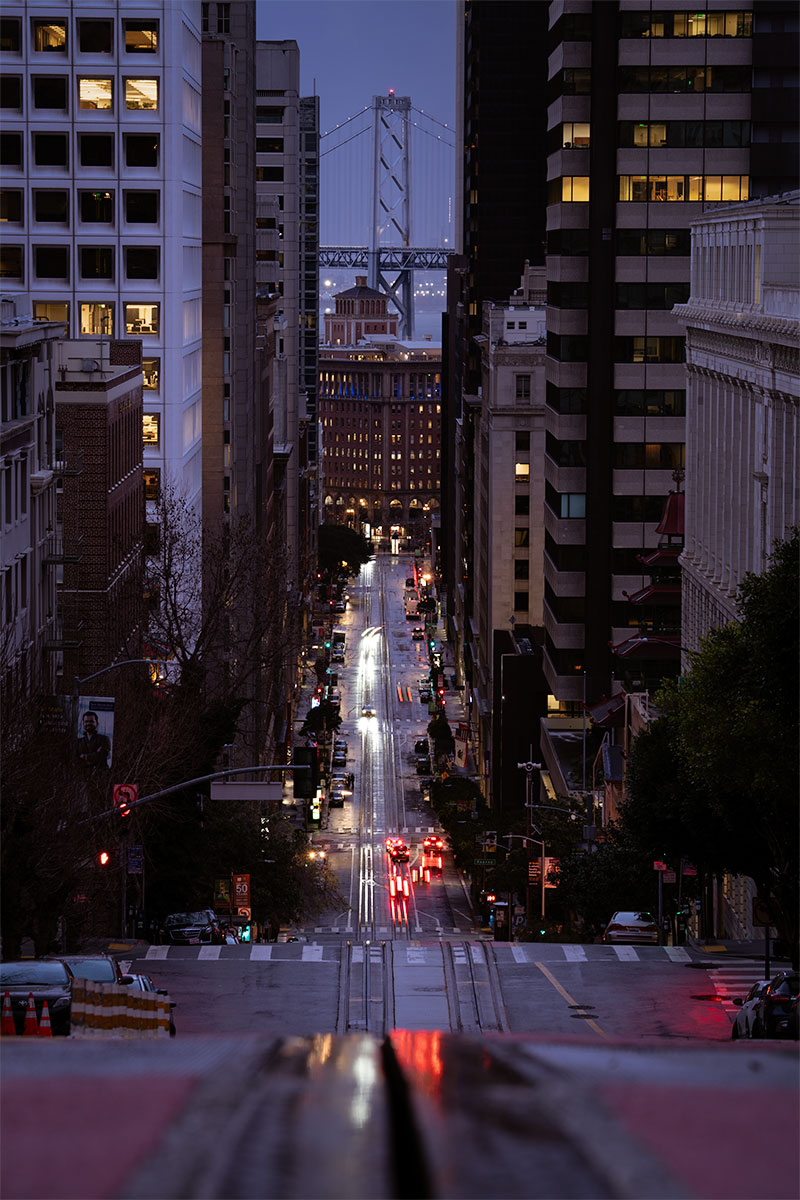 Twilight view down a San Francisco hill with cable car tracks, streetlights, and the Bay Bridge between downtown buildings